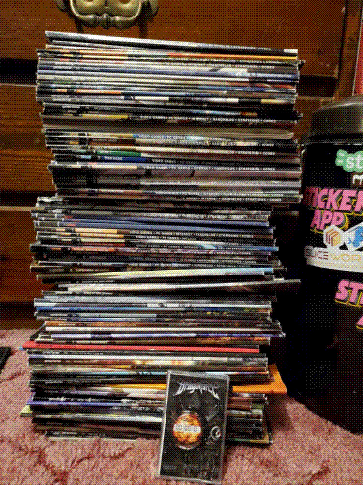 A tall stack of Game Informer magazines. They are unorganized and unevenly lined up. Next to them is a Dragonforce cassette (Inhuman Rampage) and a trashcan to give a sense of scale. The cassette is dwarfed by the height of the stack.